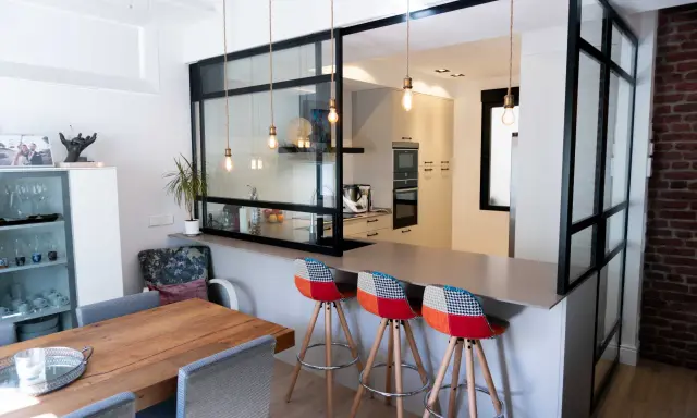 Tabique cocina semiabierta al salón con barra y taburetes coloridos. La cocina moderna se integra al salón a través de un panel de cristal y metal.