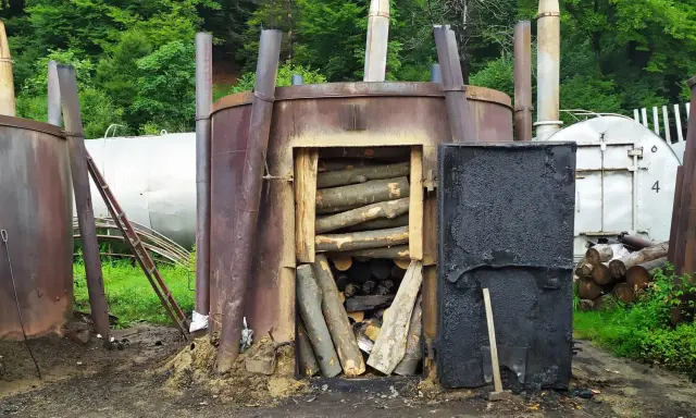 Wypalanie węgla drzewnego w piecu. Drewno ułożone w otwartym palenisku, obok czarne drzwi i inne metalowe konstrukcje.