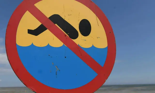 Znak zakazu kąpieli na plaży. Zamknięte kąpieliska nad morzem, mimo pięknej pogody i widocznej w oddali osoby w wodzie.