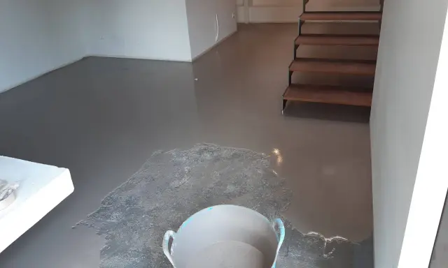 Pavimento continuo de hormigón gris recién aplicado, con una escalera de madera al fondo y un cubo con material.