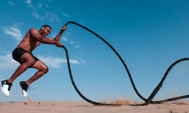 Mann mit Kopfhörern macht ein intensives HIIT-Training mit Seilen im Sand unter blauem Himmel.