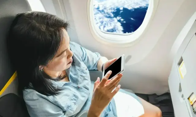 Kobieta siedzi przy oknie samolotu i patrzy na telefon.