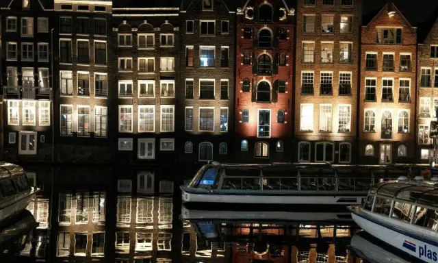 Edifici illuminati e riflessi sull'acqua, un battello turistico naviga tranquillo. Magica atmosfera di Amsterdam by night.