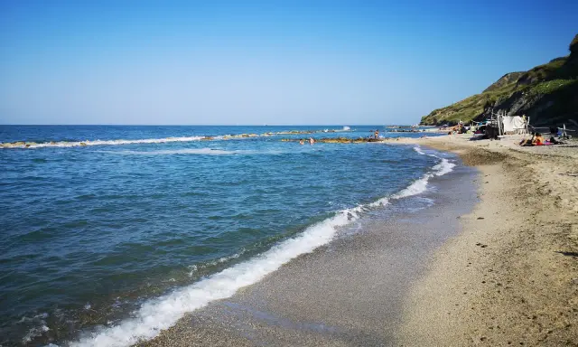 La **spiaggia** di **Castel di Mezzo** baciata dal sole, con il mare azzurro e la collina verde sullo sfondo.