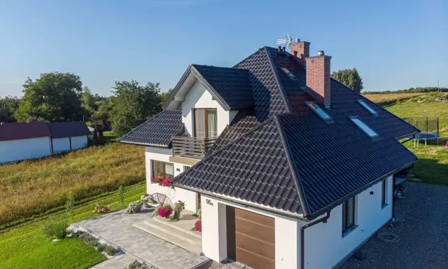 Nowoczesny dom z czarnym dachem, odpornym na warunki atmosferyczne, klasa korozyjności C4. Otoczony zielenią i zadbanym podjazdem.