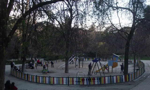 Parque de la Quinta de la Fuente del Berro con niños jugando en columpios y toboganes, rodeado de árboles y una valla colorida.