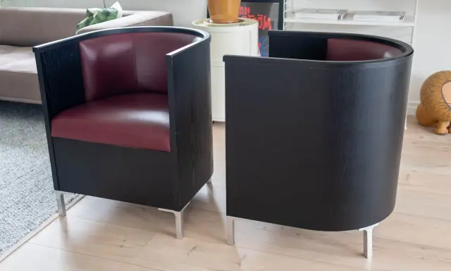 Två svarta fåtöljer med vinröda sitsar, designade av Mats Theselius, står på ett ljust trägolv.