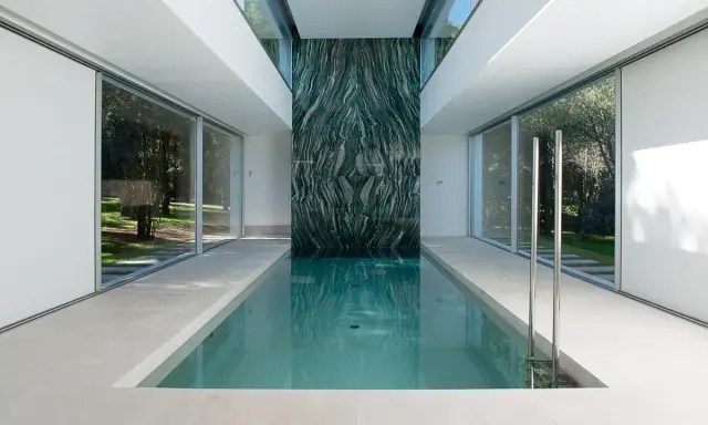Piscina interior con pared de mármol verde veteado, rodeada de ventanales que dan a un jardín.