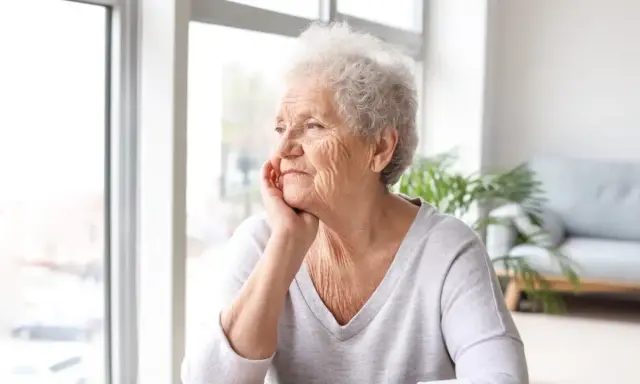 Anziana con mano sul mento, sguardo pensieroso verso la finestra. Potrebbe riflettere sulla pseudodemenza depressiva.