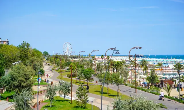 Passeggiata nel verde del parco del mare Rimini, con gente che cammina e pedala, e la ruota panoramica sullo sfondo.