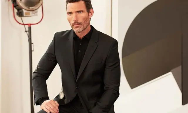 A man in a sharp black suit and polo shirt, a classic funeral outfit for men, poses with a thoughtful expression.