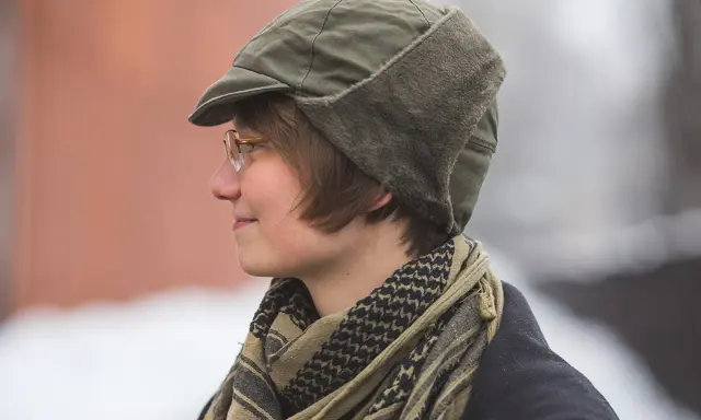 Osoba w okularach nosi zieloną czapkę polową z futrzanymi nausznikami i szalik w stylu keffiyeh.