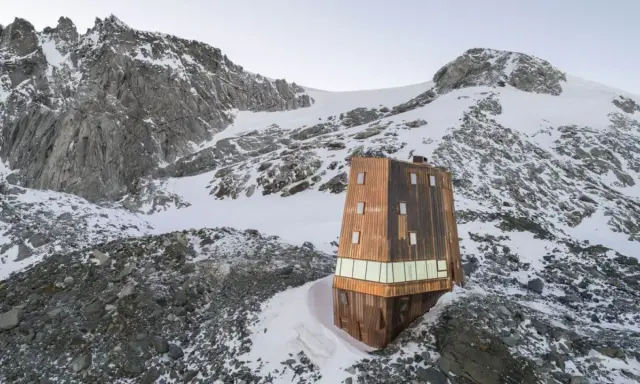 Il rifugio Sasso Nero, un'architettura moderna in legno, si erge solitario tra rocce e neve, dominato da imponenti vette montuose.