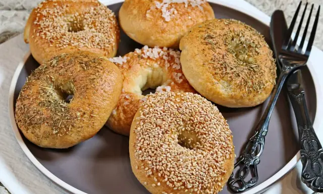 Sechs selbstgemachte Bagels auf einem Teller: zwei mit Sesam, zwei mit Kräutern und zwei mit grobem Salz bestreut. Ein Messer und eine Gabel liegen daneben.