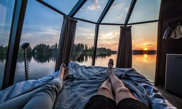 Två personer vilar i en glaskupa med utsikt över en sjö i Norrland vid solnedgången. En perfekt glampingupplevelse.