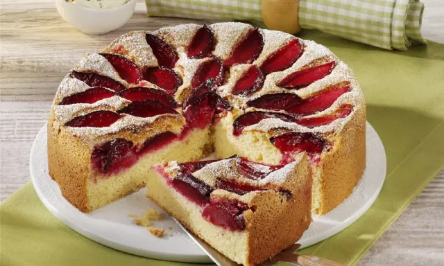 Szybkie ciasto ze śliwkami na oleju, posypane cukrem pudrem, z kawałkiem na łopatce. Obok miseczka z bitą śmietaną.