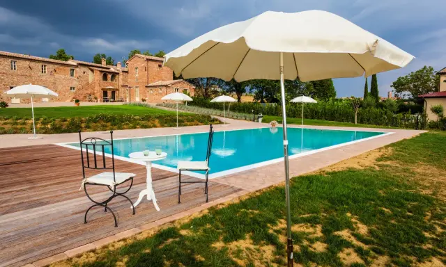 Piscina con vista su un casale in mattoni, perfetta per le tue vacanze in Toscana. Tavolino con sedie e ombrelloni pronti per rilassarsi.