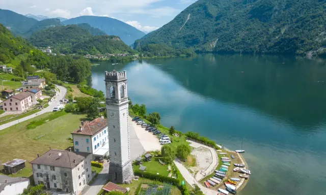 Torre campanaria affacciata sul lago di Corlo, con montagne verdi sullo sfondo e un villaggio pittoresco.