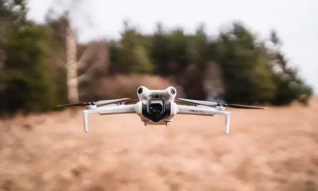 Eine weiße Drohne A3, Modell Mini 4 Pro, schwebt über trockenem Gras. Im Hintergrund verschwimmen Bäume und eine bewölkte Himmel.