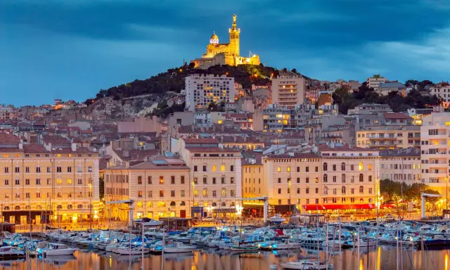 Porto di Marsiglia illuminato al tramonto, con la Basilica di Notre-Dame de la Garde in cima alla collina. Perfetto per le tue marsiglia escursioni fai da te.