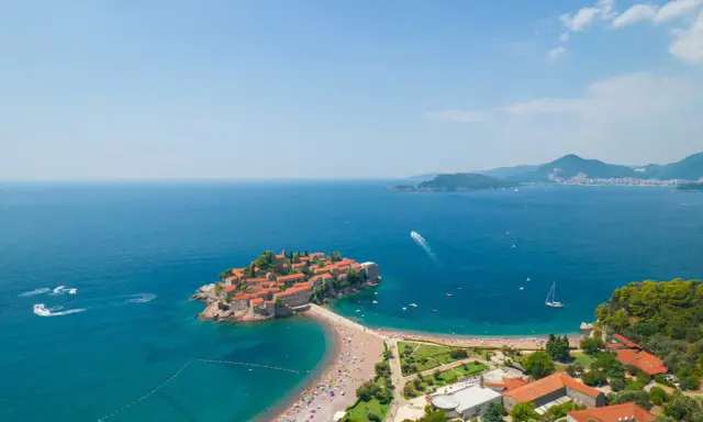 Idealna czarnogóra pogoda wrzesień: słoneczna wyspa Sveti Stefan, turkusowe morze, plaża pełna ludzi i łodzie na wodzie.