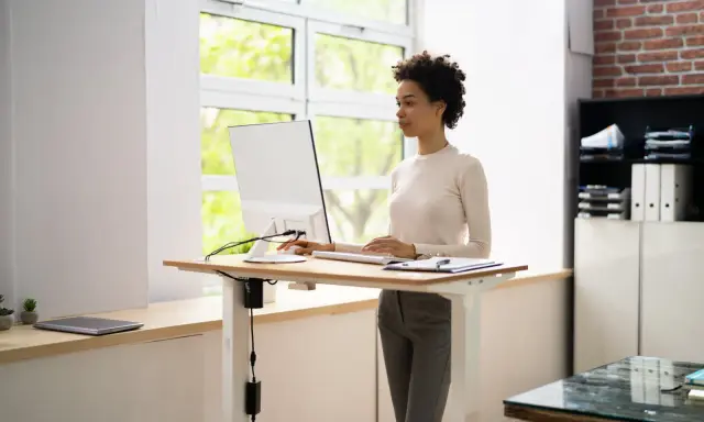 Une femme travaille sur un ordinateur à un bureau réglable en hauteur. La position debout prolongée présente des risques pour la santé, il est donc important de varier les postures.