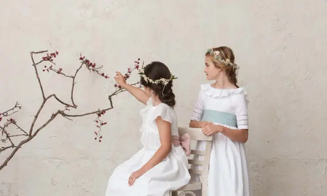 Dos niñas con vestidos blancos y coronas de flores, una sentada tocando una rama, la otra de pie. Peinados de trenza para niña de comunión.