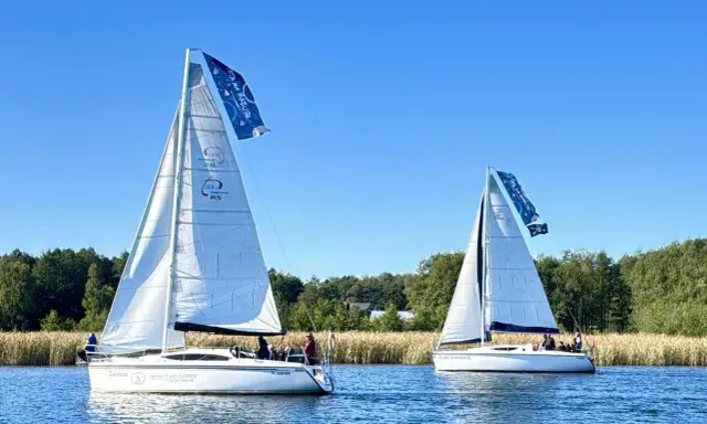 Dwa żaglowce na jeziorze, symbolizujące to, z czego słyną Mazury – żeglarstwo i piękne krajobrazy.