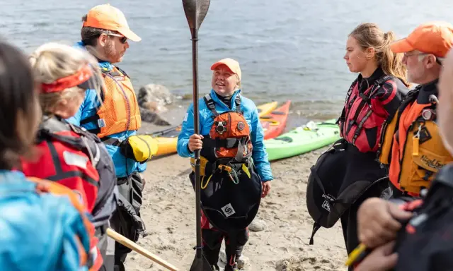 Grupp samlad på stranden, redo för paddling. En instruktör visar paddelteknik. Kajaker ligger i bakgrunden.
