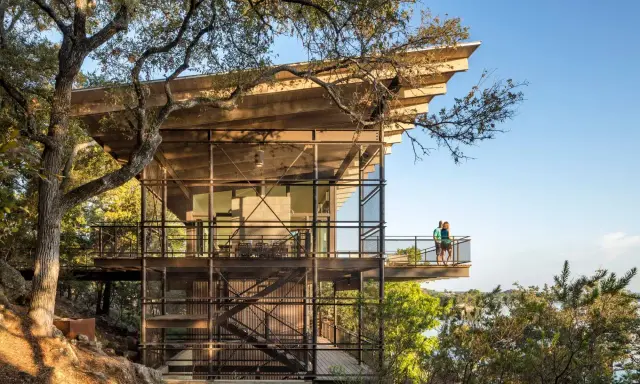Maison moderne sur un terrain en pente raide, avec des balcons et des escaliers extérieurs en métal, nichée dans les arbres près d'un lac.