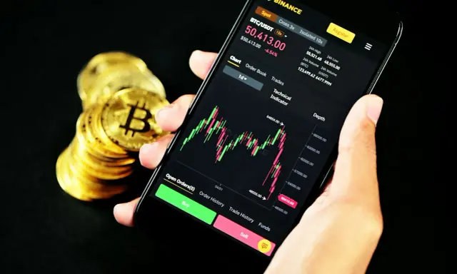 Ręka trzyma smartfon z wykresem kursu Bitcoina na platformie Binance. Obok stos złotych monet. Czy Binance czy Zonda? Decyzja należy do Ciebie.
