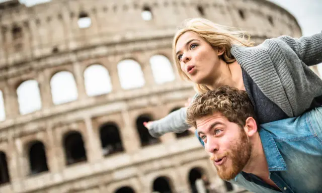Coppia in una vacanza romantica a Roma, con il Colosseo sullo sfondo. Lui porta lei sulle spalle, entrambi con espressioni sorprese.