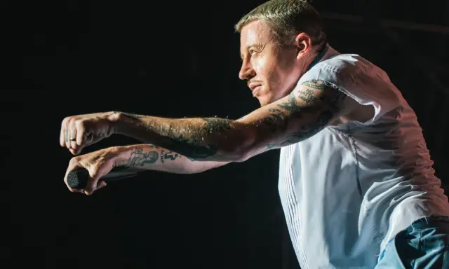 Macklemore na koncercie w Polsce, z mikrofonem w dłoni, w białej koszuli i z tatuażami na ramionach.