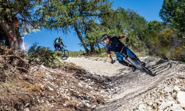 Due ciclisti affrontano un sentiero sterrato in discesa, immersi nella natura. La **tonezza del Cimone** si percepisce nell'aria fresca e nel paesaggio montano.
