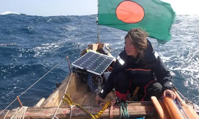 Capucine Trochet navigue sur une mer agitée, le drapeau du Bangladesh flottant au vent. Un panneau solaire est visible sur le bateau.