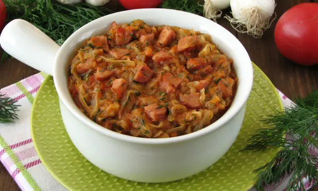 Pyszny bigos z młodej kapusty z kiełbasą, podany w białej misce na zielonym talerzyku. W tle pomidory, czosnek i koperek.