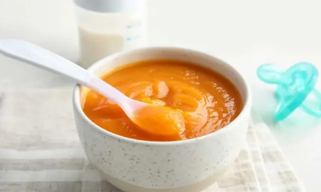Un tazón con puré de calabaza bebé, una cuchara rosa y un biberón.