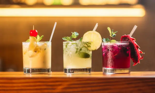 Trzy kolorowe drinki na barze: żółty z wiśnią, zielony z limonką i miętą, oraz fioletowy z pitają. Idealne do letnich wieczorów, a może i z **kazbek peak brandy z czym pić**?