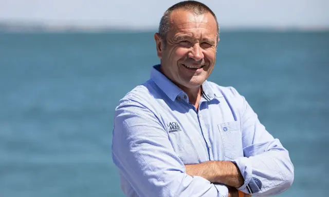 Lalou Roucayrol, souriant, bras croisés, devant la mer. Il porte une chemise bleue rayée avec un logo sur la poitrine.