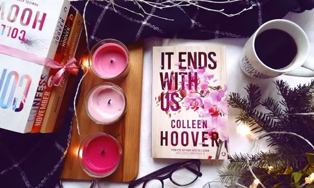 Książka "It Ends With Us" Colleen Hoover, obok świec, kawy i innych książek, tworzy przytulną atmosferę do czytania.
