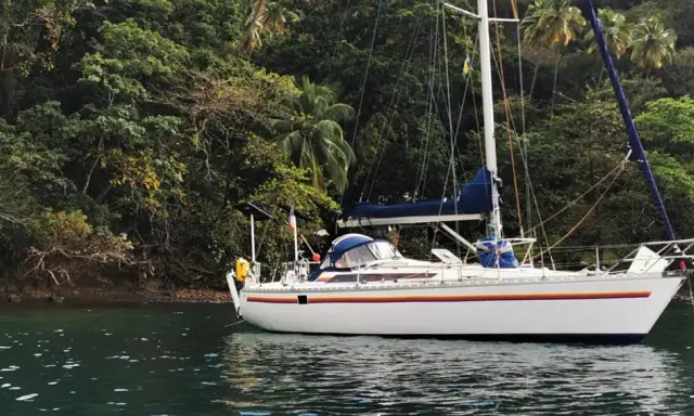 Un voilier blanc, un voilier Jurançon, est ancré dans une baie tropicale luxuriante, entouré d'une végétation dense et de palmiers.