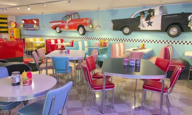Interni di un diner in **stile americano** con sedili color pastello, tavoli cromati e decorazioni a tema auto d'epoca.