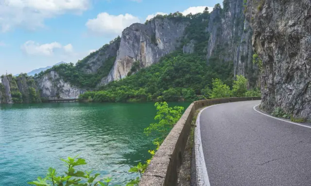 Strada panoramica lungo un lago verde smeraldo, con imponenti pareti rocciose e vegetazione lussureggiante. Un percorso suggestivo per raggiungere la baia di Bogn.