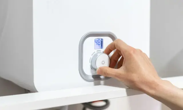 Mano ajustando el dial de una caldera para bajar la presión. La pantalla digital muestra 48°C.