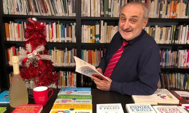 Daniele Novara, autore di libri per genitori, legge un suo libro tra scaffali pieni di volumi, con un albero di Natale e una bottiglia di spumante.