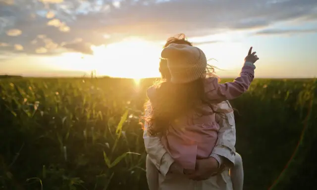 Bambina aggrappata alla schiena di un adulto, con il sole al tramonto che illumina un campo. Un'immagine di puro attaccamento affettivo.