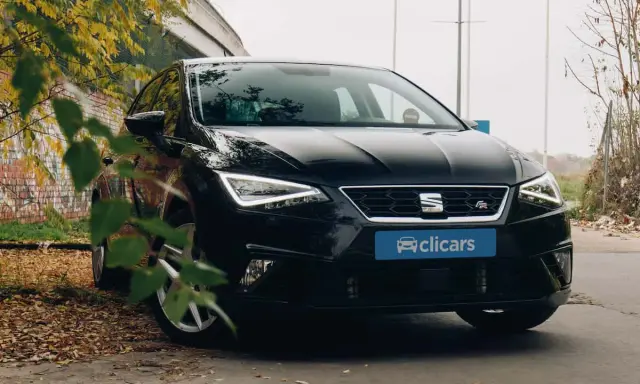 Coche negro SEAT León FR aparcado entre hojas secas y un muro de ladrillos. El emblema "FR" indica su versión deportiva.