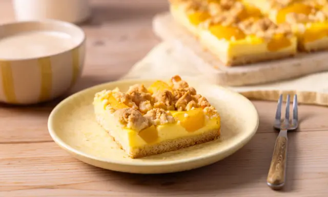 Ein Stück saftiger Pfirsich Blechkuchen mit Vanillepudding und Streuseln auf einem gelben Teller, daneben eine Gabel.