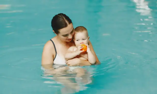 Mama trzyma roczne dziecko na basenie. Maluch bawi się pomarańczową piłeczką.