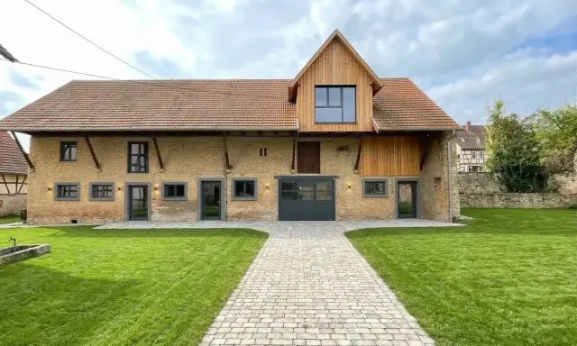 Belle grange rénovée avec une façade en briques jaunes, un toit en tuiles et un ajout moderne en bois. Un chemin pavé mène à l'entrée principale.